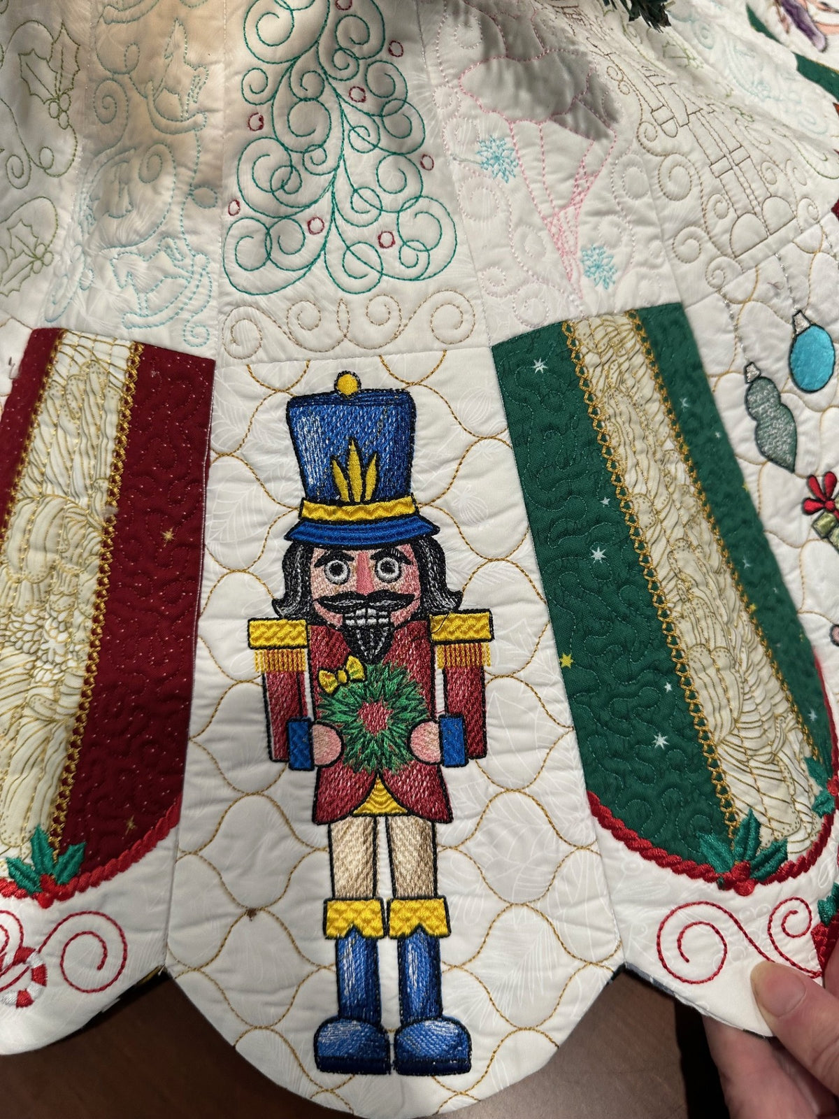 Detail view of a handcrafted white Christmas tree skirt featuring detailed embroidered Nutcracker Ballet characters around the border with detailed quilting in the center. Close up of Nutcracker.
