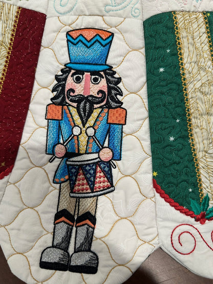 Detail view of a handcrafted white Christmas tree skirt featuring detailed embroidered Nutcracker Ballet characters around the border with detailed quilting in the center. Close up of Nutcracker.
